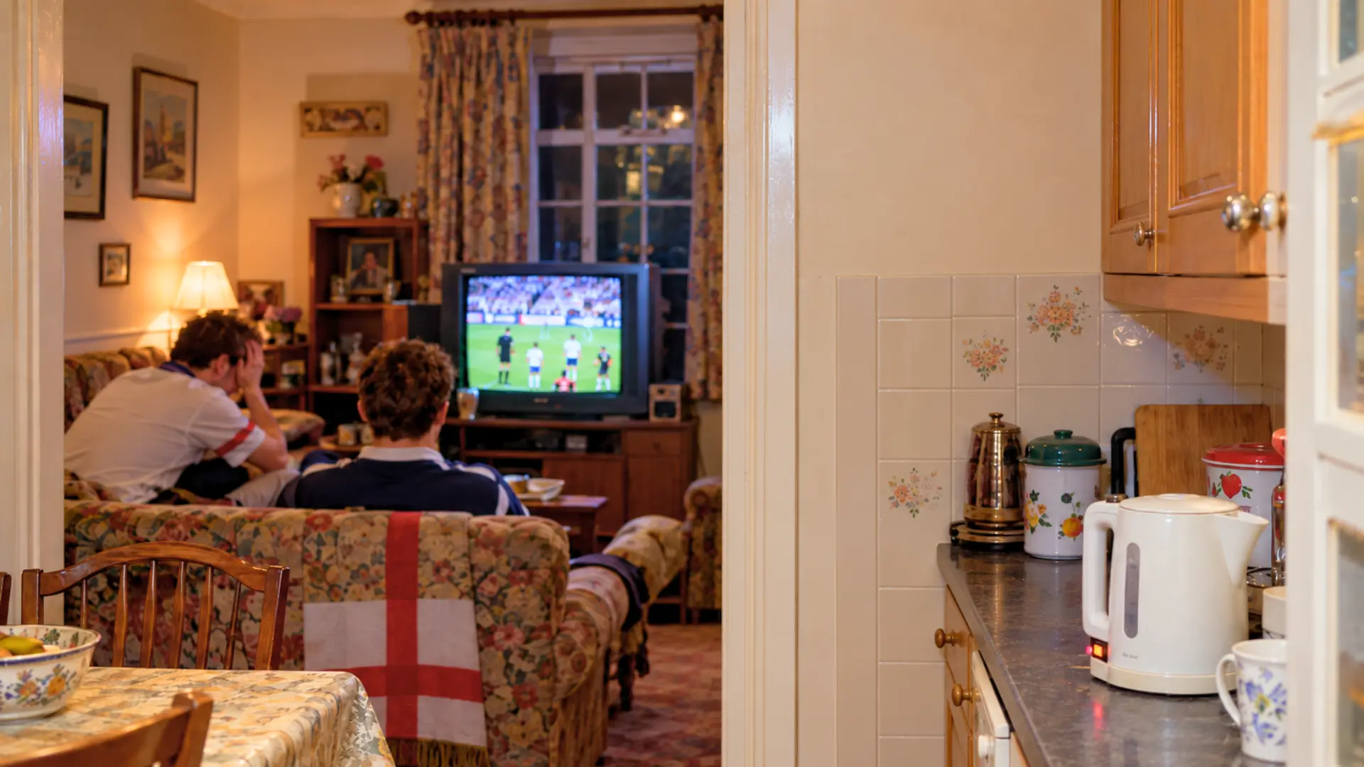 Bouilloire électrique allumée dans une cuisine britannique de 1990, avec deux supporters anglais déçus devant un match de football à la télévision.